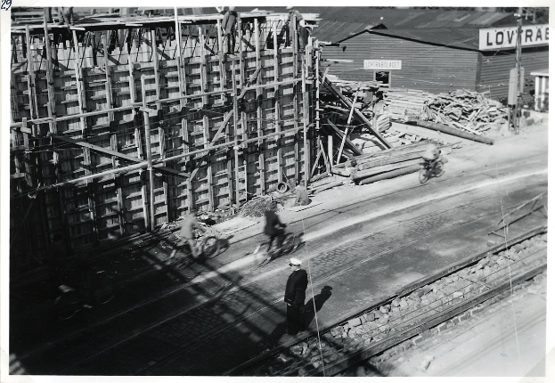 Schwarz-weiß-Foto einer Baustelle mit einem teilweise errichteten Gebäude, Menschen auf Fahrrädern und zu Fuß auf der Straße, Bahnschienen, Pfosten, Bretter und andere Gegenstände, mit Text unten.
