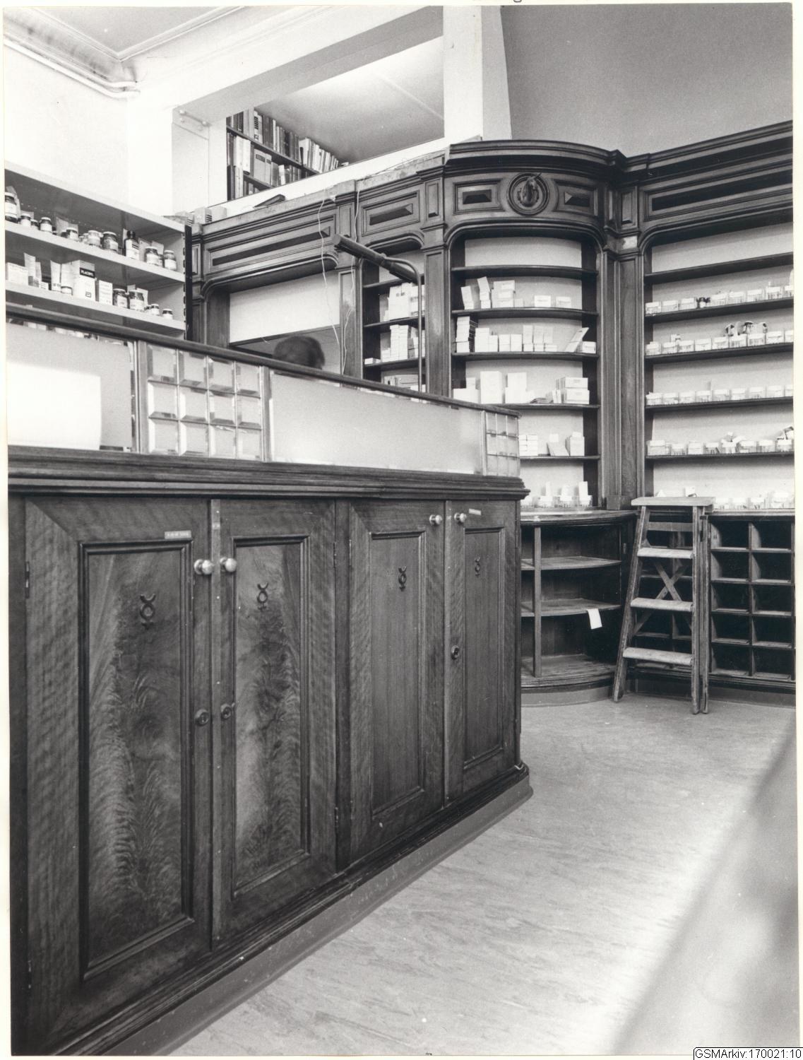 Schwarzes und wei├čes Foto eines vintage Apothekeninneren mit einer Theke, Schüben, einer Leiter, Regalen mit Beh├Ąltern und Text überall.