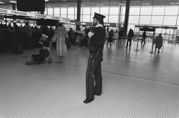 Ein Polizist in Uniform und Mütze steht in einem Flughafen-Terminal und hält eine Waffe, umgeben von Menschen und Gepäckwagen, Stangen, Texttafeln, Deckenleuchten und Glaswänden im Hintergrund.