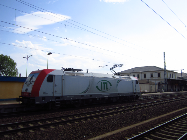 Eine diesel-elektrische Lokomotive fährt auf Schienen neben einem Gebäude, mit Strommasten, Kabeln, Straßenlaternen, Bäumen und einem bewölkten Himmel im Hintergrund.