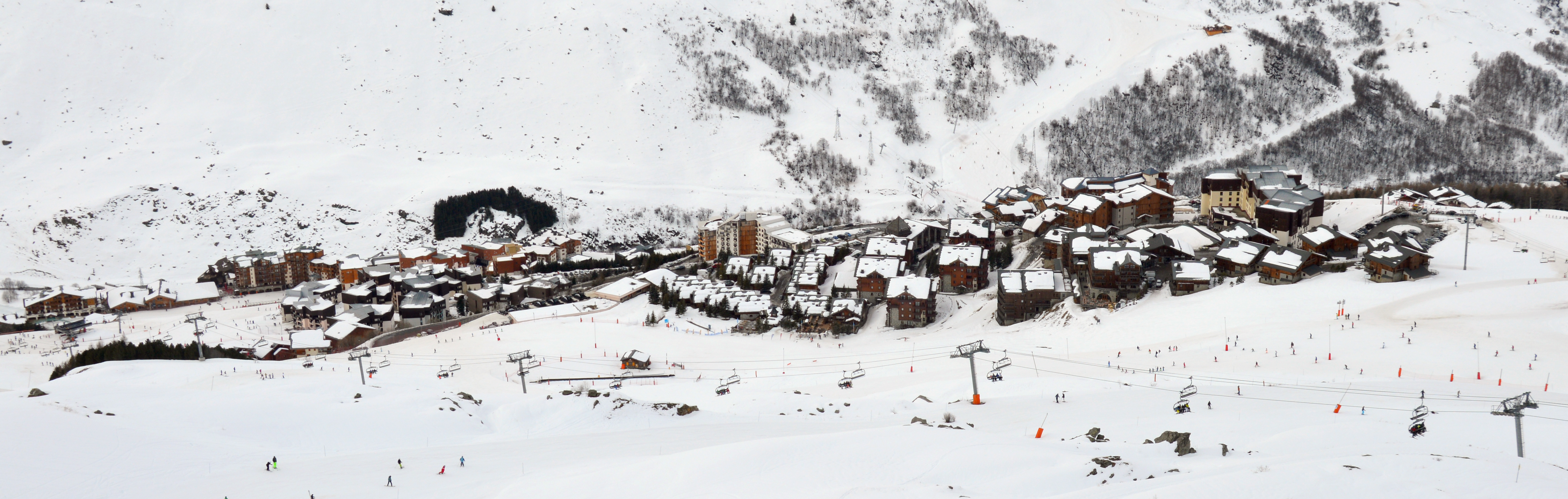 Ein kleines Dorf liegt in einer schneebedeckten Berglandschaft, mit Häusern, Strommasten, Drähten und Bäumen, während einige Menschen eine Piste hinunterskifahren.