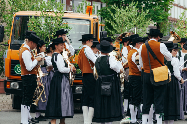 Gruppe von Menschen in traditioneller bayrischer Tracht, die eine Straße entlangmarschieren, Musikinstrumente spielen und Taschen tragen, mit Bäumen und Gebäuden im Hintergrund und einem geparkten Fahrzeug.