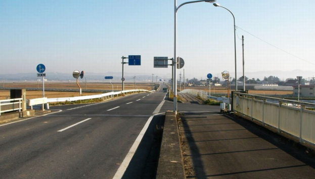 Eine Autobahnüberführung mit Geländern, Laternenmästen und Verkehrszeichen, mit Gebäuden, Bäumen, Hügeln und einem klaren blauen Himmel im Hintergrund.