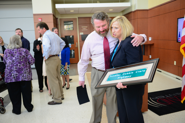 Eine Gruppe von Menschen, die sich drinnen aufhalten, wobei einer ein Foto mit der Aufschrift 'Senator Debbie Clary Highway' hält, mit einer Flagge, einer Wand, einer Tür und Lichtern im Hintergrund.