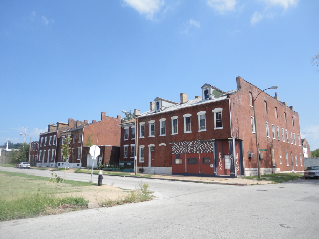 Ein Gebäude mit Masten und Straßenlampen, umgeben von Bäumen und Fahrzeugen auf beiden Seiten, mit einem Weg, Gras und einem Feuerlöscher unten und Himmel oben.