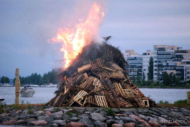 Holzobjekte brennen mit Feuer, Steine unten, Wasser in der Mitte mit einem Schiff darauf, Gebäude, Bäume und Himmel im Hintergrund.