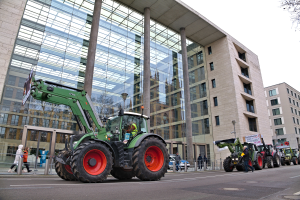 Eine Gruppe von Traktoren fährt auf einer Straße vor einem Gebäude, mit Menschen auf dem Gehweg und einem Baum auf der rechten Seite, unter einem sichtbaren Himmel, wahrscheinlich an einer Demonstration teilnehmend.