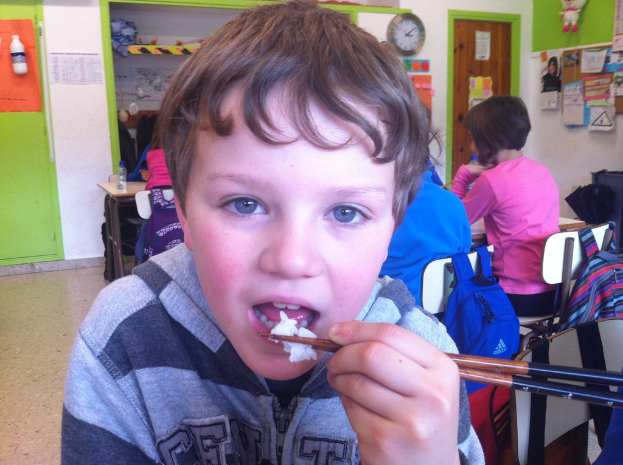 Ein Junge in einer grauen und schwarzen Jacke isst mit Stäbchen in einem Klassenzimmer, mit anderen Kindern hinter ihm, einer grün-weißen Wand, Lehrplakaten an einer weißen Wand, einer braunen Tür und einer Wanduhr.