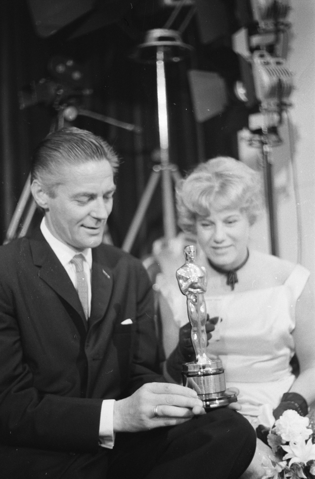 Ein Mann in einem schwarzen Blazer, weißem Hemd und Krawatte hält eine Oscar-Statue, während er neben einer Frau in Abendgarderobe sitzt, die einen Blumenstrauß hält, beide von Hintergrundlichtern in einem Schwarz-Weiß-Bild beleuchtet.