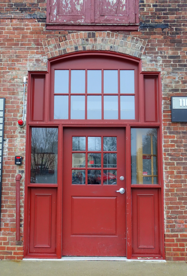 Eine rote Brandschutztür an einem Backsteinbau mit Glasfenstern, einer Türklinke und einer nummerierten Tafel auf der linken Seite.