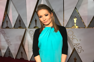 Eine Frau in einem blauen und schwarzen Kleid steht auf einem roten Teppich vor einer Tafel mit der Aufschrift "Oscars 2018 Red Carpet Arrivals" und verschiedenen Skulpturen im Hintergrund.