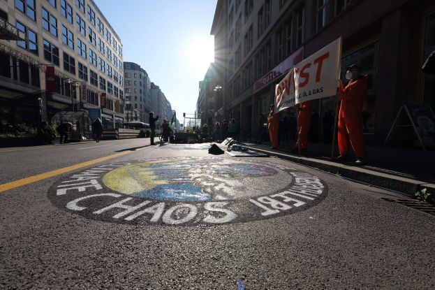 Eine Gruppe von Menschen mit Protestschildern und Transparenten auf einer Stadtstraße mit Gebäuden, Pflanzen und einer gemalten "Chaos" auf der Straße.