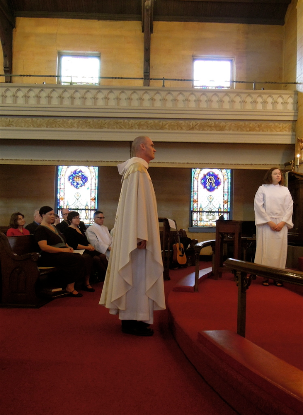 Eine Frau und ein Mann in weißen Kleidern stehen in einer Kirche, mit einem roten Teppich zu ihren Füßen, Bänken links, Musikinstrumenten im Hintergrund und Geländern, Fenstern und Wänden im gesamten Bild.
