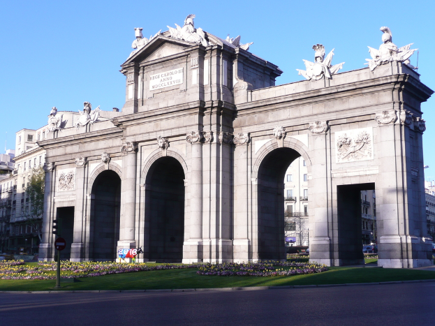Gebäude und Skulpturen in der Mitte, mit einer Straße, Gras, Blumen, einem Pfahl und einer Platte unten und Himmel oben.
