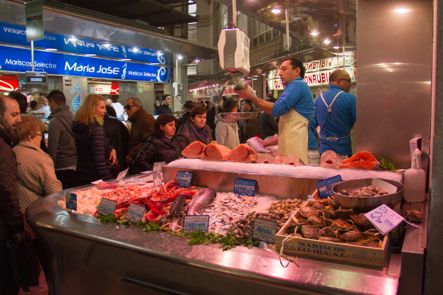 Eine Marktszene mit Menschen, die gehen und einige Fische schneiden, mit Fischen und Brettern unten und Geschäften, Brettern, Pfählen und Lichtern im Hintergrund.