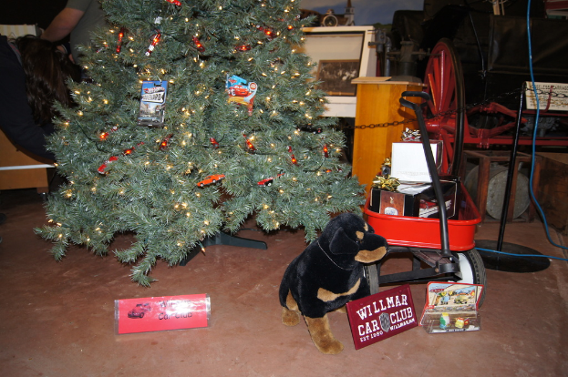 Ein Hundespielzeug, Namensschilder, ein Einkaufswagen mit Gegenständen, ein Weihnachtsbaum, ein Rad, ein Pfahl, eine Kette, eine Holzplatte, eine stehende Person und verschiedene andere Gegenstände im Hintergrund.