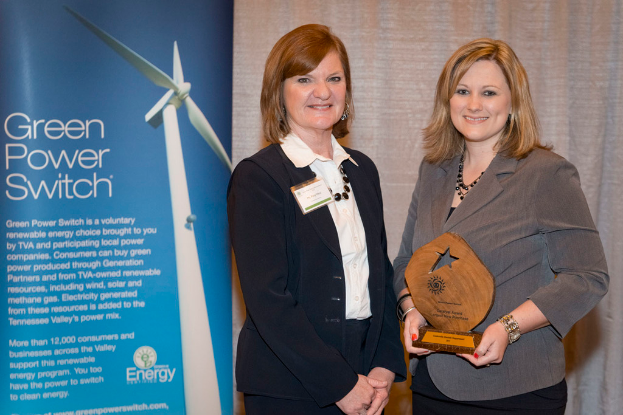 Zwei Frauen stehen in einem Raum, eine hält einen Preis, mit einem Banner neben ihr und einem Vorhang im Hintergrund.