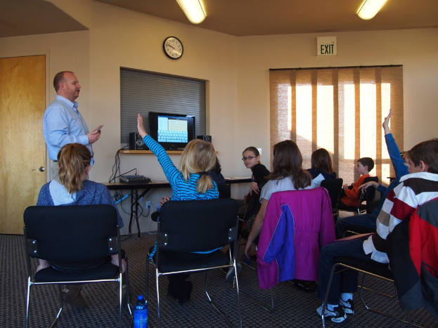 Ein Mann steht in einem Klassenzimmer und spricht zu einer Gruppe sitzender Kinder, wobei ein Monitor, Lautsprecher, eine Uhr, ein Fenster und eine Tür im Hintergrund zu sehen sind.