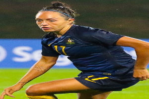 Eine Fußballspielerin im Einsatz auf dem Feld, die ein blaues T-Shirt und gelbe Shorts trägt, mit einem unscharfen Hintergrund, der ein blaues Schild mit Text zeigt.