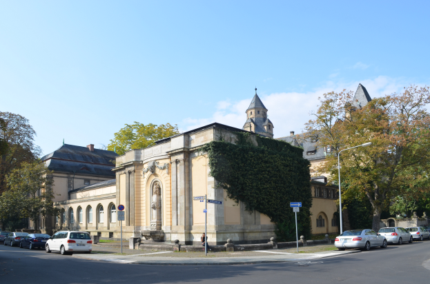 Die Kirche St. Peter und St. Paul in Heidelberg, Deutschland, ein großes Gebäude mit einem zentralen Glockenturm, umgeben von Bäumen, Straßenlaternen, Autos, Fußgängern und Schildern, unter einem bewölkten Himmel.