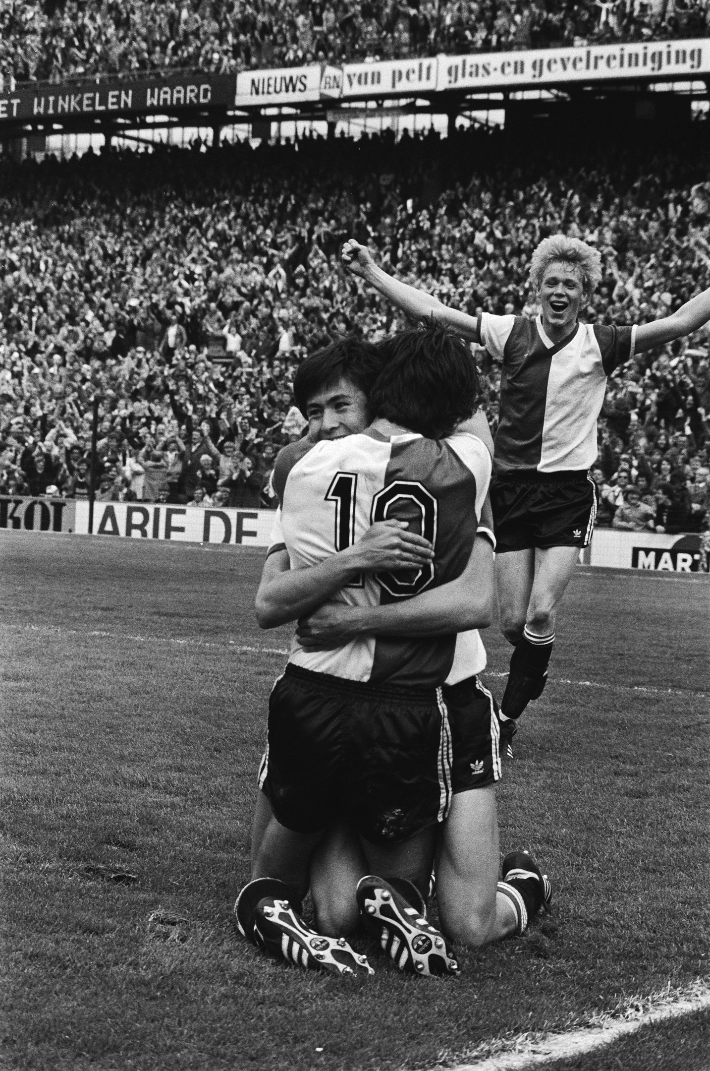 Drei Fußballspieler umarmen sich auf dem Boden nach einem Tor, mit einer jubelnden Menge und schwenkenden Fahnen im Hintergrund, in einer schwarz-weißen Stadionatmosphäre.