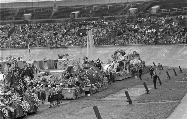 Schwarzes und weißes Foto einer Stadionparade mit Menschen, die stehen und sitzen, einem zentralen Springbrunnen und Blumengebinden auf Fahrzeugen.