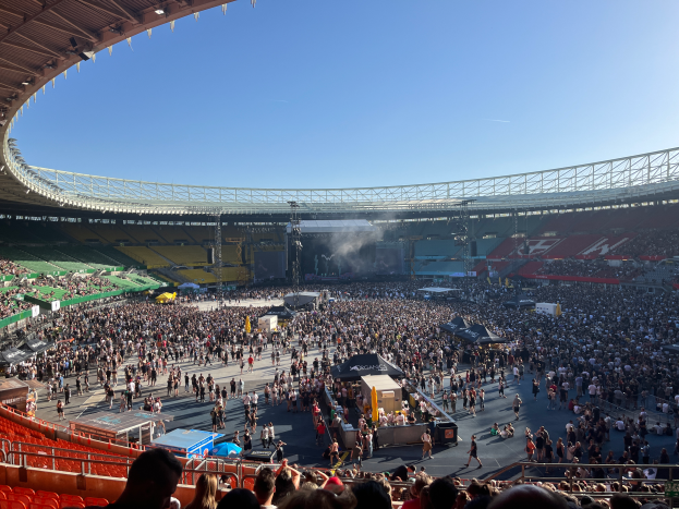 Eine große Menschenmenge versammelt sich in einem Stadion, mit einer Bühne in der Mitte, auf der verschiedene Gegenstände zu sehen sind, und einem Dach mit Lichtern oben, das ein Konzert im Wembley-Stadion vermuten lässt.