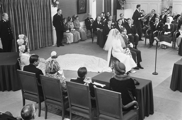 Schwarzes und weißes Foto einer Hochzeitszeremonie mit sitzenden und stehenden Gästen, einem Mann mit Mikrofon, einer Frau mit Blumenstrauß, einem Podium mit Mikrofon, Blumenschmuck, einem Vorhang, einem gerahmten Foto an der Wand und einer Tür im Hintergrund.