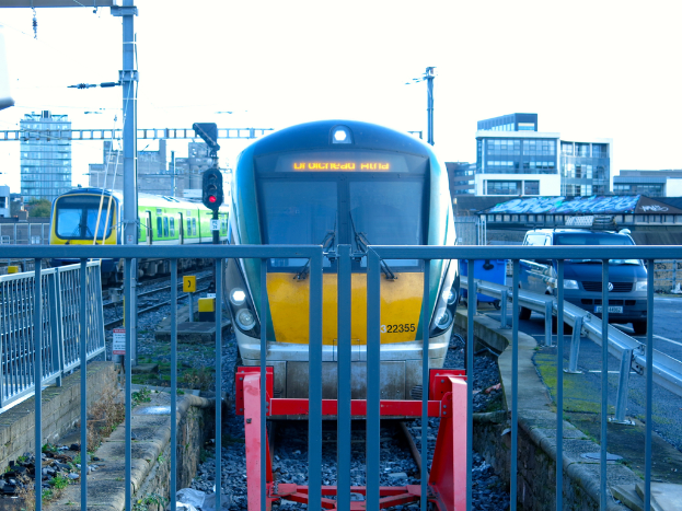 Zwei Züge auf Schienen mit einem Zaun im Vordergrund, Pfosten mit einem, der ein Signallicht hat, ein Auto auf der Straße rechts, Gebäude im Hintergrund und Himmel oben.