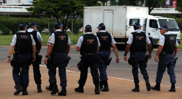 Eine Gruppe von uniformierten Beamten, darunter Mützen, Hemden, Schutzwesten, Uhren, Hosen und Schuhe, die auf einer Straße mit einem geparkten Lkw auf der rechten Seite neben einem Zaun, Gebäuden, Bäumen, Pflanzen, Masten und Schildern im Hintergrund und einer Brücke oben rechts gehen.