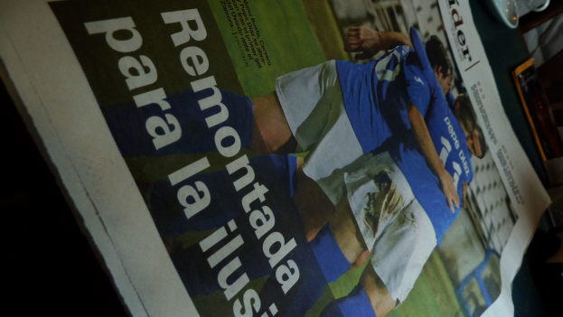 Eine Zeitung auf einem Tisch zeigt drei Männer in blauen T-Shirts, möglicherweise beim Fussballspielen, mit Text unten im Bild.