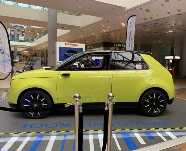 Ein gelbes wasserstoffbetriebenes Elektroauto vor einem Gebäude geparkt, umgeben von Absperrungen und Bannern, mit Menschen in der Nähe und einer Topfpflanze.