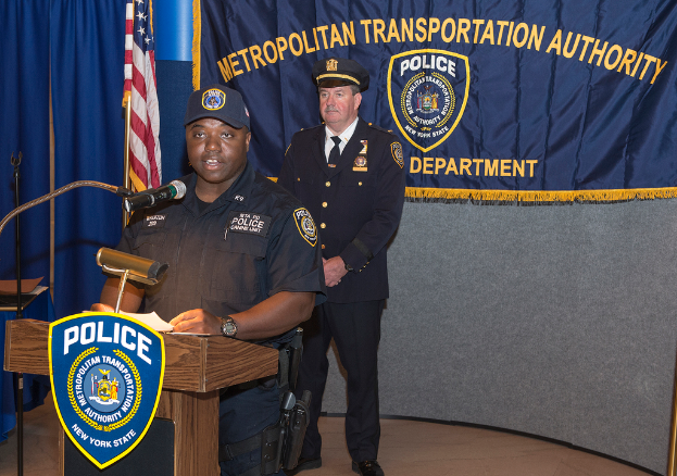 Zwei Polizeibeamte stehen vor einem Rednerpult, einer hält Papiere und spricht in ein Mikrofon, mit einer Tafel, Flagge und Tisch in der Nähe, und einer Tafel mit Symbolen und Text im Hintergrund.