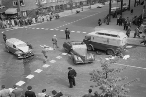 Eine Schwarz-Weiß-Szene eines Autounfalls an einer Straßenkreuzung, mit mehreren Fahrzeugen einschließlich eines Lieferwagens, einer Gruppe von Menschen, die herumstehen, und Gebäuden, Bäumen, Pfählen und Texttafeln im Hintergrund.