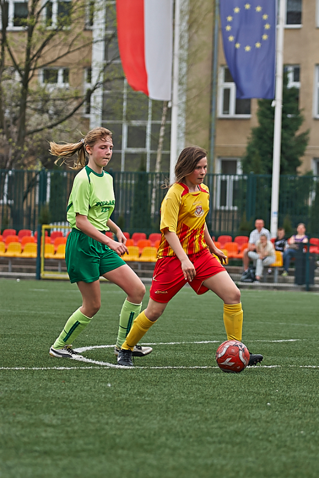 Zwei Frauen spielen Fußball auf einem Feld mit einem Ball, während im Hintergrund vier Personen auf Stühlen sitzen und ein Gebäude sowie Fahnen zu sehen sind.