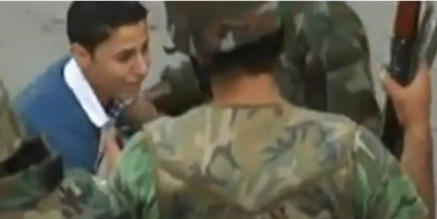 Ein Militär-Offizier in der Mitte hält eine Waffe, mit einem Jungen vor ihm und einem weiteren Militär-Offizier links daneben.