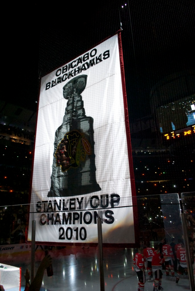 Eine nächtliche Stadionkulisse mit einem großen Banner in der Mitte, Zuschauern im Hintergrund, einem Glas auf dem Boden und einigen Spielern auf dem Feld, beleuchtet von Stadionlampen.