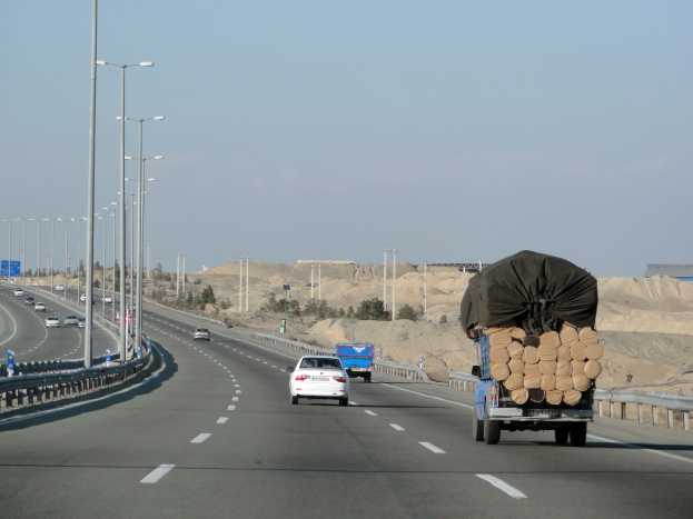 Ein Lkw mit einer großen Ladung Holz fährt auf einer Autobahn, umgeben von Leitplanken, Laternen, Texttafeln, Bäumen und Sand, mit Hügeln und einem klaren blauen Himmel im Hintergrund.