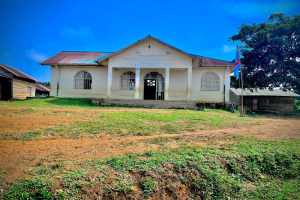 Ein kleines weißes Gebäude mit einer Fahne oben drauf, umgeben von Gras und Bäumen, unter einem klaren blauen Himmel, mit Fenstern, Türen und Text an der Wand, der darauf hinweist, dass es eine Schule für Kinder ist.