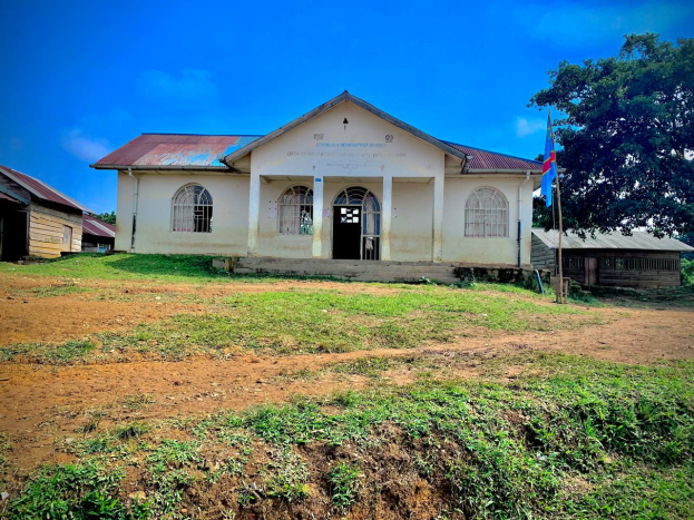 Ein kleines weißes Gebäude mit einer Fahne oben drauf, umgeben von Gras und Bäumen, unter einem klaren blauen Himmel, mit Fenstern, Türen und Text an der Wand, der darauf hinweist, dass es eine Schule für Kinder ist.