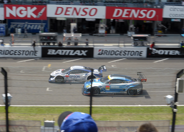 Zwei Sportwagen rasen auf einer Rennstrecke mit einer Zuschauermenge hinter einem Maschendrahtzaun, Polen und Lichtern, mit Texttafeln im Hintergrund.