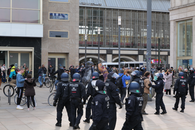Eine Gruppe von Polizisten in Helmen steht vor einer Menge, die Fahrräder hält, mit Glaswänden, Laternen und Fahrzeugen im Hintergrund.