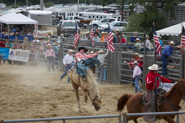 Zwei Personen zu Pferd nehmen an einem Charreada-Event teil, umgeben von Zuschauern, hängenden Bannern und Fahnen, aufgestellten Zeltplanen, parkenden Fahrzeugen und Bäumen im Hintergrund.