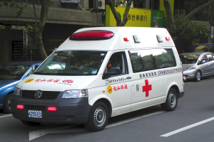 Ein Rettungswagen steht am Straßenrand geparkt, umgeben von anderen Fahrzeugen, mit Bäumen, Gebäuden, Texttafeln und Klimaanlagen im Hintergrund.