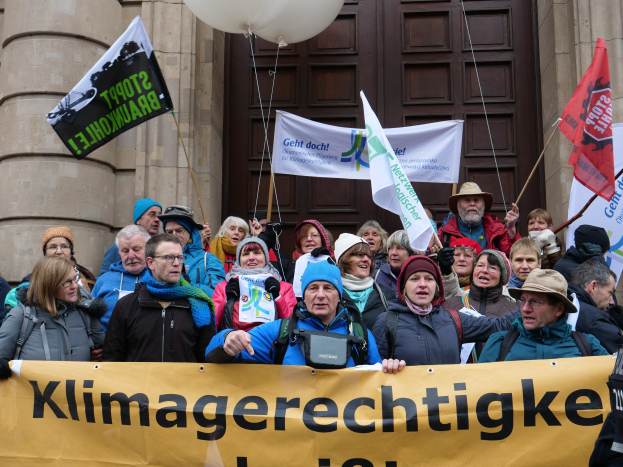 Eine Gruppe von Menschen vor einem Gebäude mit Säulen und einer Tür, die eine Fahne mit Text, Flaggen mit Stangen und Luftballons halten, nimmt an einem Klimastreik in Deutschland teil.