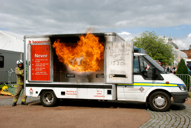 Ein brennender Transporter mit Schrift darauf ist im Vordergrund, dahinter steht ein Mann. Im Hintergrund sind Gebäude, einige Menschen, Pflanzen, ein Baum und ein bewölkter Himmel zu sehen.