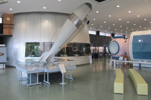 Ein geräumiger Raum mit verschiedenen Weltraumausrüstungen, darunter ein rechts stehendes Raket, Sofas auf dem Boden, eine Tafel mit Text auf einem Tisch, ein Zaun, eine Wand, ein Dach mit Deckenlampen und ein Fenster.