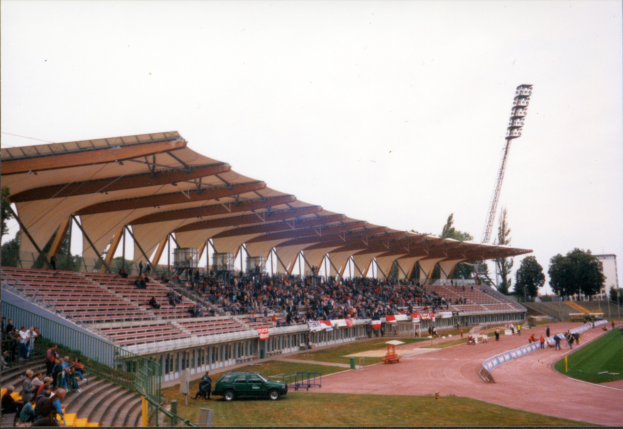 Altes Schwarz-Weiß-Foto eines Stadions voller Zuschauer, das sitzende Fans unter der Dach zeigt, ein geparktes Auto auf dem Feld, stehende Besucher auf dem Rasen, Stützpfähle und Metallrahmen, Umzäunung, Straßenpfahl, entfernte Bäume, hoher Turm und bewölkter Himmel.