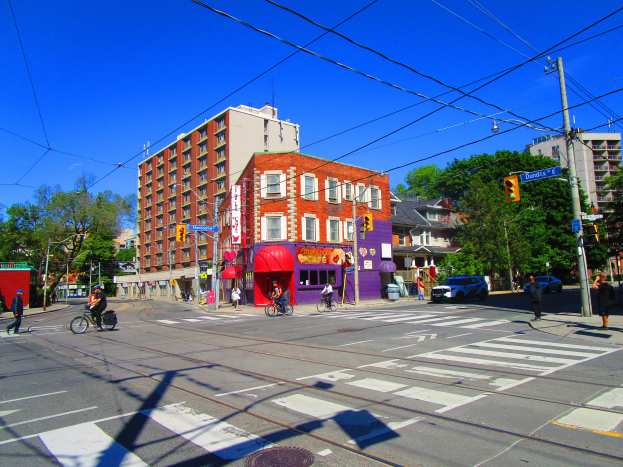 Eine belebte Stadtstraße mit Menschen, die an einer Ampel über die Straße gehen, Fahrzeugen, Radfahrern, Gebäuden, Bäumen, Laternen, Verkehrszeichen und Texttafeln unter einem sichtbaren Himmel.