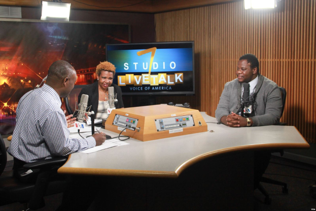 Zwei Männer und eine Frau sitzen an einem Tisch in einem Studio und führen ein Live-Gespräch mit Papieren und Mikrofonen vor sich, einem Fernseher und einem Bildschirm im Hintergrund.